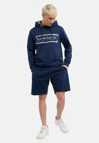 Navyblauwe hoodie met een voorzak, voorzien van witte logo-tekst. Gecombineerd met bijpassende shorts, beide in een zachte stof. Witte sneakers maken de look compleet.