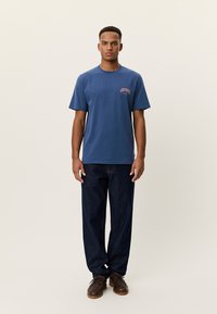 Μπλε βαμβακερό t-shirt με στρογγυλή λαιμόκοψη και κοντά μανίκια, με πορτοκαλί λογότυπο στο αριστερό μέρος· συνδυασμένο με σκούρο μπλε τζιν παντελόνι.