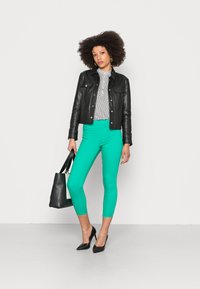 Casaco de cabedal preto, blusa branca com bolas, leggins verdes brilhantes e tacões altos pretos. Bolsa tote preta com um detalhe em branco.