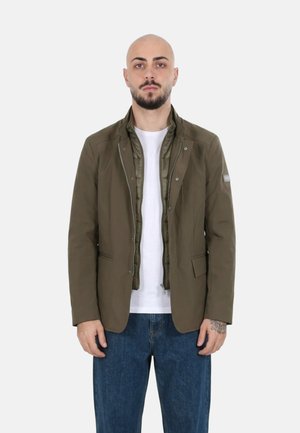 Uomo con barba e tatuaggio che indossa una giacca verde oliva a strati sopra una camicia bianca e jeans blu, in piedi su uno sfondo semplice.