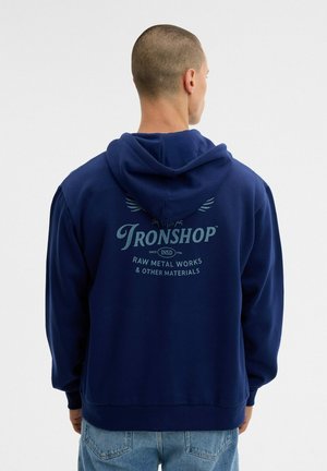 Un hombre visto de espaldas lleva una sudadera con capucha azul marino con el texto «Ironshop» y unos vaqueros azul claro, sobre un fondo blanco liso.