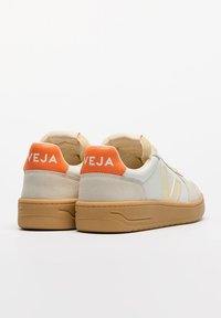 Zapatillas blancas y beige con acentos naranjas, que presentan un diseño de ante texturizado y cuero liso, y una suela de goma con un dibujo de banda vertical.