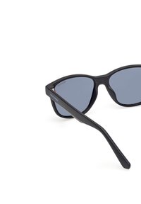 Occhiali da sole neri polarizzati con design angolare, lenti grigio scuro e finitura opaca sulla montatura. Presentano un astuccio slanciato.