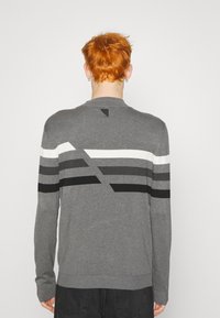 Sudadera gris de manga larga, con franjas diagonales en negro, blanco y gris en la espalda, tela de punto y puños acanalados.