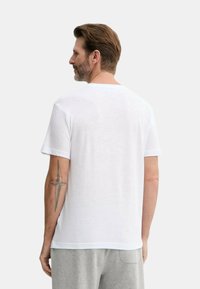 Maglietta bianca a maniche corte con colletto rotondo e design semplice, realizzata in un materiale morbido. Tatuaggio visibile sul braccio destro.