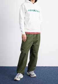 Felpa bianca con testo e logo "JORDAN" verdi, sovrapposta a una maglietta rossa. Pantaloni verde oliva e sneakers grigie con dettagli bianchi.