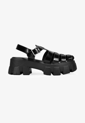 Sandalias negras de tacón ancho con diseño de parte superior trenzada, que cuentan con una correa con hebilla y suela de goma gruesa con formas de bloque distintivas.