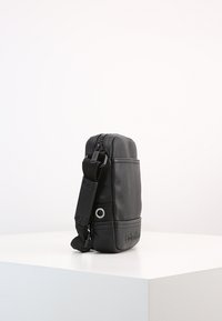 Borsa crossbody in pelle nera con finitura testurizzata, chiusura con zip, tracolla regolabile e dettaglio in metallo a forma circolare. Forma rettangolare compatta.