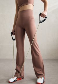 Brune, høytlivsbukser laget av elastisk stoff, kombinert med hvite og røde joggesko. Personen holder motstandsband med svarte håndtak.