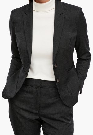 Svart texturerad blazer med skräddarsydd passform, slagkrage, två knappar och matchande byxor. Bärs över en vit polotröja.