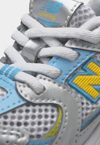 Κοντινή λήψη λευκών και ανοιχτό μπλε κορδονιών και διχτυού παπουτσιού με κίτρινο λογότυπο New Balance και την επιγραφή "running" στην ετικέτα της γλώσσας.