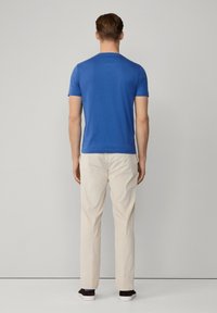 Niebieska krótkorękawowa koszulka widziana z tyłu, zestawiona z beżowymi spodniami i ciemnym obuwiem. Prosty fason, standardowe dopasowanie, minimalne detale.