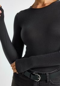 Maglietta nera a maniche lunghe con un design aderente, realizzata in tessuto a costine. Presenta aperture per i pollici nei polsini ed è abbinata a una cintura nera.