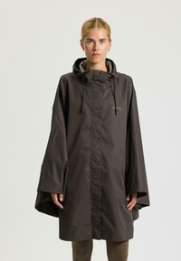 Donna che indossa un poncho impermeabile con cappuccio di colore marrone scuro, dotato di bottoni a pressione e maniche ampie, in piedi davanti a uno sfondo chiaro e semplice.