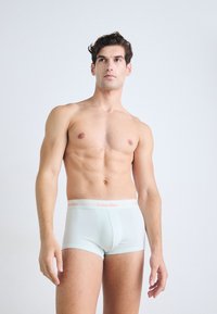 Spierkrachtige man staat rechtop, draagt lichtblauwe Calvin Klein boxershorts en kijkt licht omhoog tegen een egale witte achtergrond.
