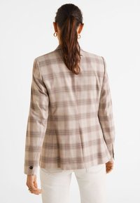 Žena s hnědým ohonem, oblečená v béžovém kostkovaném blejzru a bílých kalhotách, stojící čelem k nám proti jednoduchému bílému pozadí.