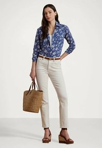 Γυναίκα ντυμένη με μπλε φλοράλ πουκάμισο, λευκά παντελόνια, καφέ ζώνη και πλατφόρμες, κρατώντας μια πλεκτή σακούλα από ψάθα, σε στάση μπροστά από φplain φόντο.