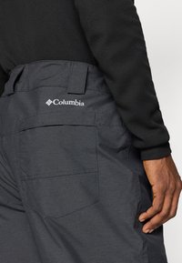 Tumedat hallid Columbia püksid tekstuurse kangaga, küljes tagatasku ja logo, kombineeritud musta pika varrukaga t-särgiga.