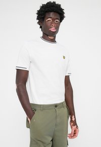 T-shirt en coton blanc avec des bordures grises au col et aux manches, arborant un petit logo jaune. Associé à un pantalon cargo vert olive.
