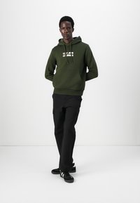 Dark green cotton hoodie with a front pocket and logo, paired with black cargo pants and black sneakers featuring white stripes.