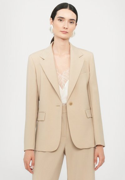 Beige maatwerk blazer met gestructureerde schouders, enkele knoopsluiting en dubbele voorkruiden. Soepele stoftextuur.