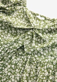 Zelena cvjetna haljina s malim bijelim cvjetovima na teksturiranom materijalu. Ima nabore detalje i nabranu struk za opušteni kroj.