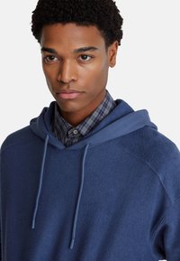 Marineblauwe hooded sweatshirt met textuurstof, trekkoord en een geruite shirtkraag eronder. Close-up van de halslijn en borst.