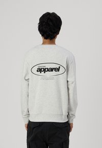 Šviesiai pilkas džemperis su ilgomis rankovėmis, apvalia iškirpte ir juodu grafiniu spausdiniu nugaros pusėje, kuriame yra užrašas "apparel" ir prekės ženklo detalės.