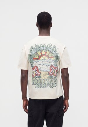 Uomo di spalle, indossa una maglietta color crema con sole colorato, onde, fiori e logo “Hollister Surf Co” sulla schiena, sfondo semplice.