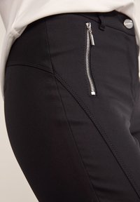 Zwarte broek met een zilveren ritszak, een gladde textuur en schuine stiksels voor een aansluitende uitstraling.