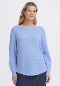 Femme aux cheveux blonds portant des boucles d'oreilles argentées en forme d'anneau et un pull en tricot bleu clair, debout devant un fond blanc uni.