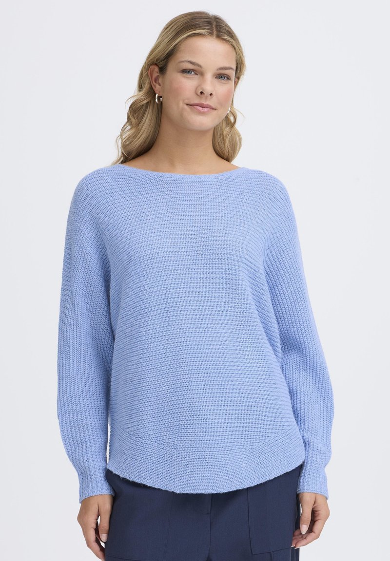 Femme aux cheveux blonds portant des boucles d'oreilles argentées en forme d'anneau et un pull en tricot bleu clair, debout devant un fond blanc uni.