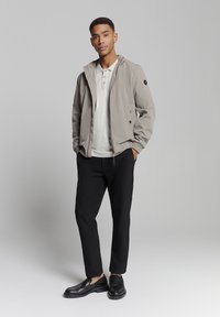 Lichtgrijze bomberjack met capuchon, met ritssluiting, zwarte accenten, gedragen over een lichtgrijs poloshirt en zwarte tapered pants, gecombineerd met zwarte instappers.