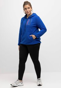 Sudadera de felpa azul cobalto con un bolsillo en el pecho con cremallera, detalles en negro y bolsillo canguro; acompañada de leggings negros y zapatillas grises.