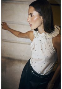 Mouwloze beige top met zwarte stippen en een rimpelcollar, gecombineerd met gestructureerde zwarte broek en een brede riem.