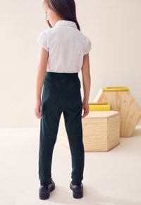 Camicia bianca con colletto e maniche arrotolate, abbinata a pantaloni verde scuro. Scarpe nere con suole spesse. Design semplice e su misura.