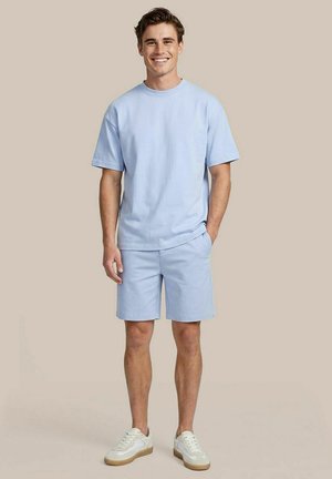 Jonge man lacht, draagt een lichtblauw T-shirt met korte mouwen en bijpassende korte broek met witte sneakers, staand tegen een beige achtergrond.
