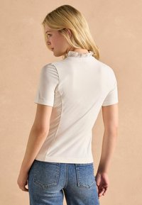 Top alb cu m�âneci scurte, detaliu de guler volanat, design ajustat și țesătură netedă, asortat cu blugi din denim, într-o croială confortabilă.