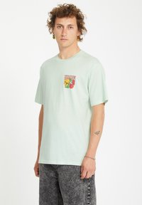 Camiseta de algodón verde claro con mangas cortas y cuello redondo. Presenta un pequeño gráfico colorido en el pecho con acentos florales.