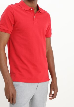 Punainen puuvillapolo-paita lyhyillä hihoilla, klassinen kaulus ja kolmen napin lista. Yhdistetty vaaleanharmaisiin housuihin.