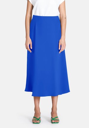 Frau trägt einen leuchtend blauen Midirock, weißes kurzärmeliges Oberteil und grüne Riemchensandalen, steht vor einem einfarbigen Hintergrund.