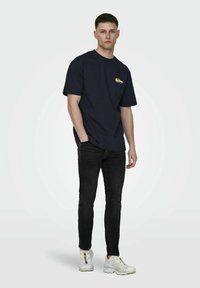 Mørkeblå t-skjorte med korte ermer og et lite gult grafisk motiv på brystet, kombinert med sorte jeans og hvite sneakers. Enkel design og passform.