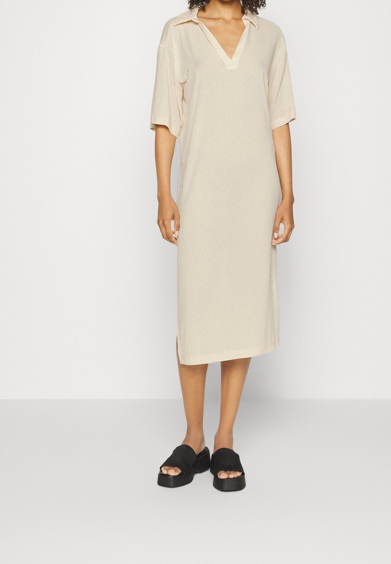 Robe beige longueur genou avec décolleté en V et manches courtes. Texture douce, fentes latérales, associée à des sandales noires à larges brides.