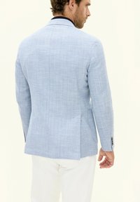 Blazer blu a spina di pesce con revers a notched, spacco singolo, due tasche laterali e dettaglio del colletto in contrasto blu navy. Indossato con pantaloni chiari.