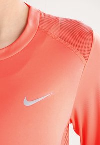 Nike Sportshirt in leuchtendem Korallenrot mit glatter Textur und Mesh-Akzenten an der Schulter. Mit silbernem Logo auf der Brust.