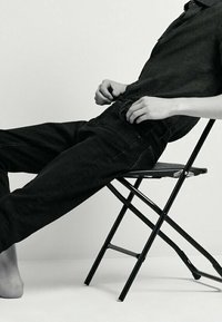 Svarta denimjeans med avslappnad passform och synlig sömnad, tillsammans med en mörk skjorta, sittande på en svart vikstol.