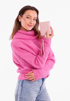 Pink knitted sweater with a high cowl neck, paired with light blue denim. Holding a small pink wallet with a wrist strap and metallic accents.