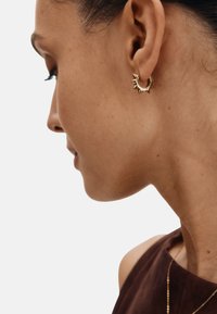 Guldørering i form af en hoop med spidse accenter, tekstureret overflade, båret i højre øre, placeret mod en neutral baggrund.