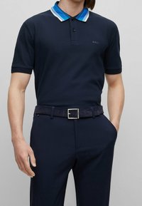 Marinblå polo med en blårandig krage, kortärmad och med en diskret logotyp. Tillsammans med mörka byxor och ett mockabälte.