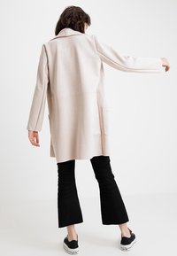 Donna vista da dietro che indossa un lungo cappotto beige chiaro, pantaloni neri a vita alta e sneakers nere con suola bianca.
